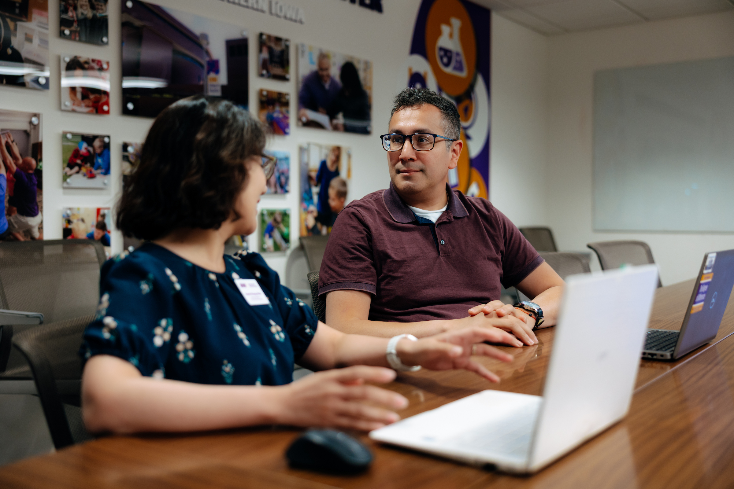 Two people sit across from each other at a conference table in a meeting room, engaged in discussion. Open laptop computers are positioned on the table in the foreground. One participant gestures while speaking, while the other looks toward them. The room contains several office chairs and a wall display featuring photographs and graphics related to campus or community activities. A large whiteboard is mounted on the wall in the background, and the space is arranged for collaboration, planning, or professional meetings.