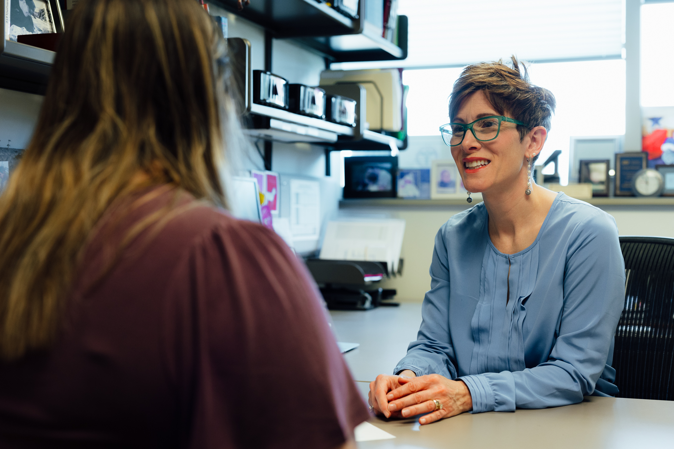 Two people sit across from each other at a desk in an office. One person, seated behind the desk, wears glasses and a light blue blouse and is engaged in conversation with the other person, who is visible from behind. Office shelves, framed photos, documents, and organizational supplies are visible in the background, with daylight coming through a window. The setting suggests a one-on-one meeting, advising session, or consultation.