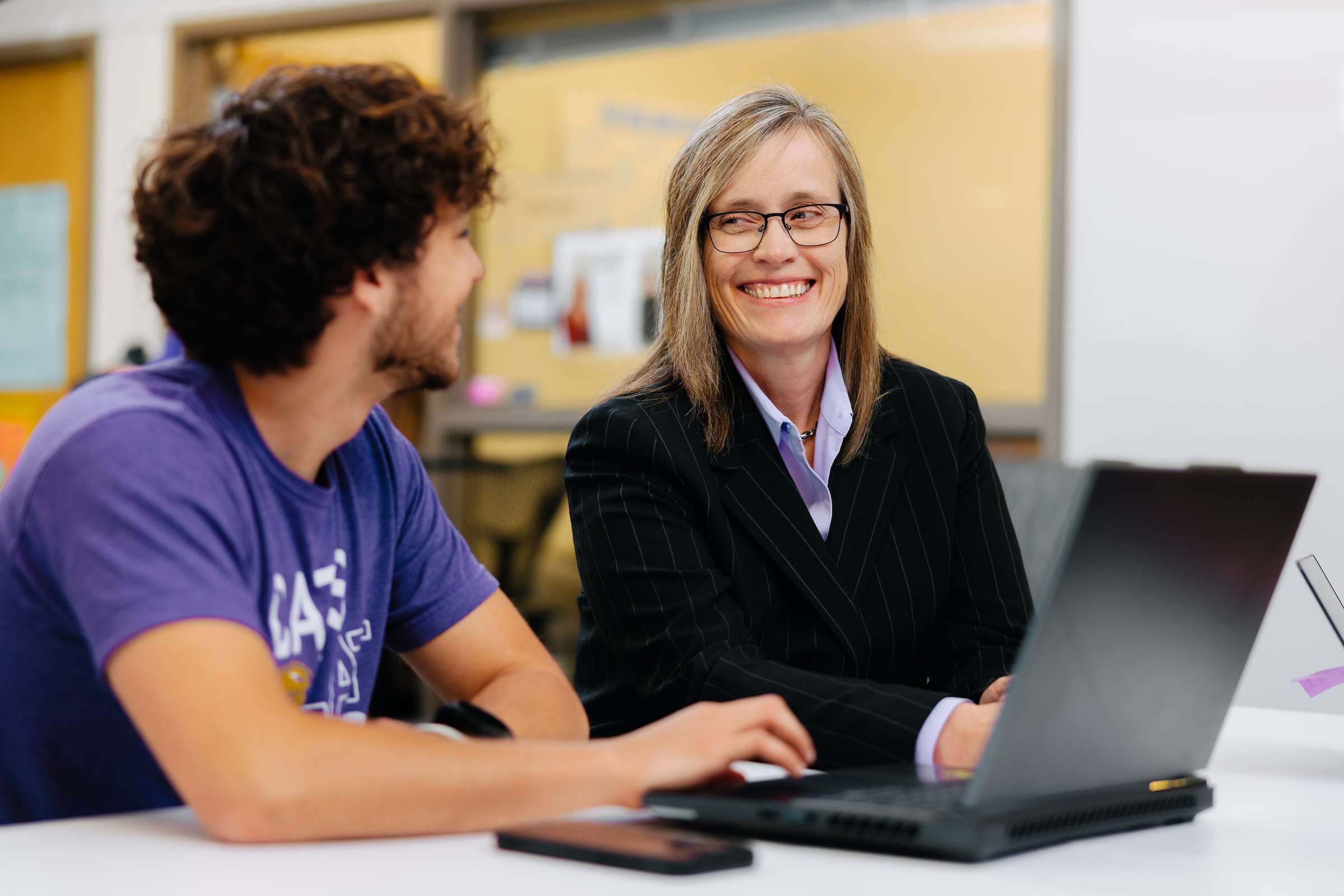 Two people sit together at a table in a modern learning or office environment, working on a laptop computer. One person wearing a purple UNI T-shirt sits in the foreground with a laptop and smartphone on the table. Across the table, another person wearing glasses, a dark pinstriped blazer, and a light-colored collared shirt sits beside the laptop. Glass-walled rooms and office spaces are visible in the background. The image captures a one-on-one conversation or mentoring interaction in a professional and co