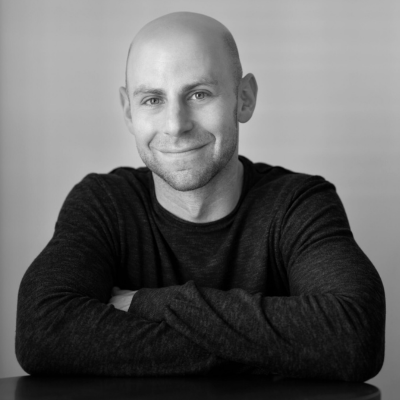 Black-and-white portrait of a Adam Grant wearing a dark long-sleeve shirt, seated at a table with his arms crossed. He is smiling gently and looking directly at the camera against a plain, softly lit background.