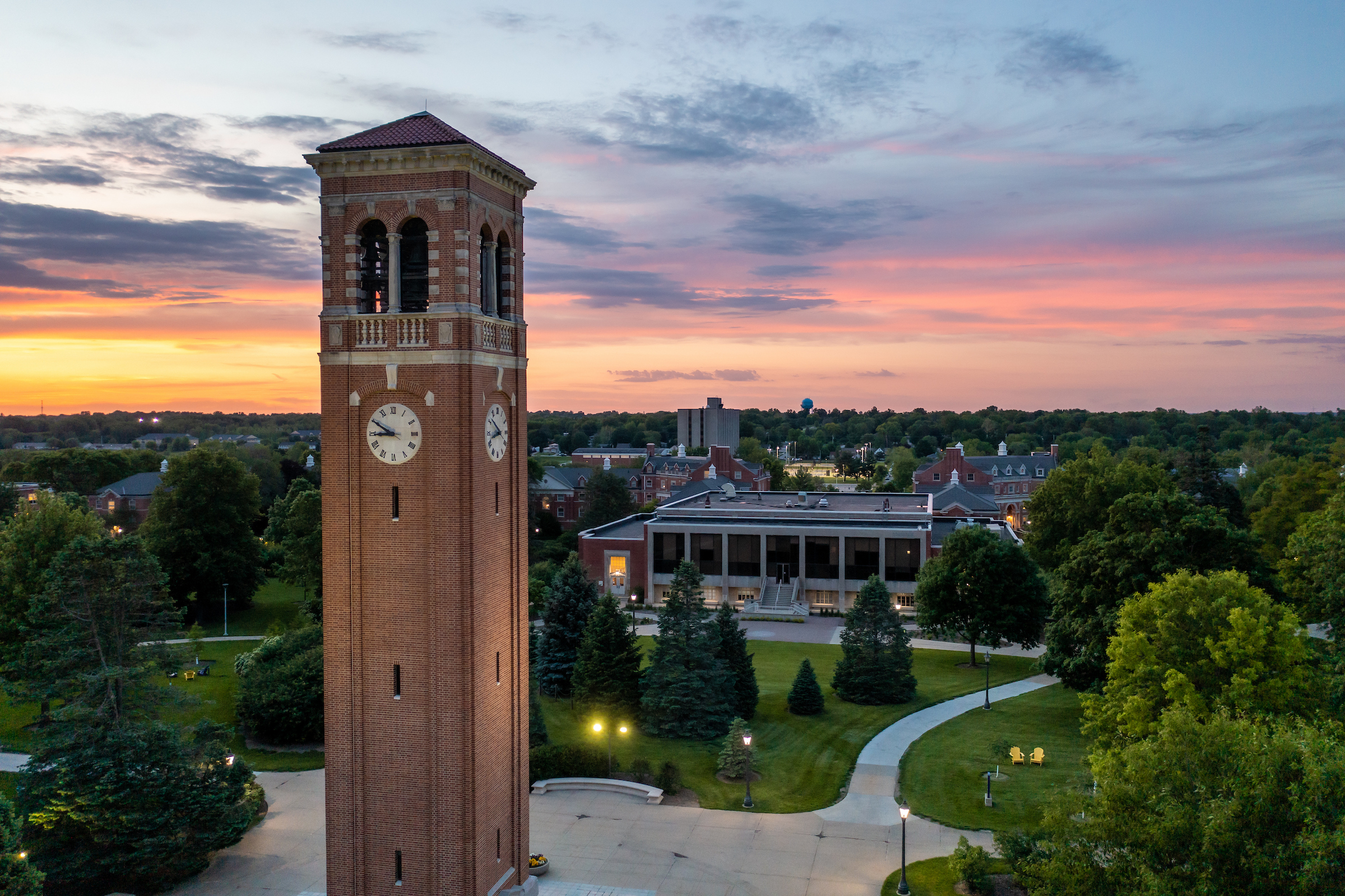 Aerial view of a university campus at sunset featuring a tall brick clock tower with clock faces visible on multiple sides. The tower rises above landscaped grounds with winding walkways, mature trees, and surrounding academic buildings. The sky is filled with pink, orange, blue, and purple hues from the setting sun, casting soft light across the campus. Building lights are beginning to illuminate the scene, and the tree-lined campus extends into the distance toward the horizon. The image highlights the clo