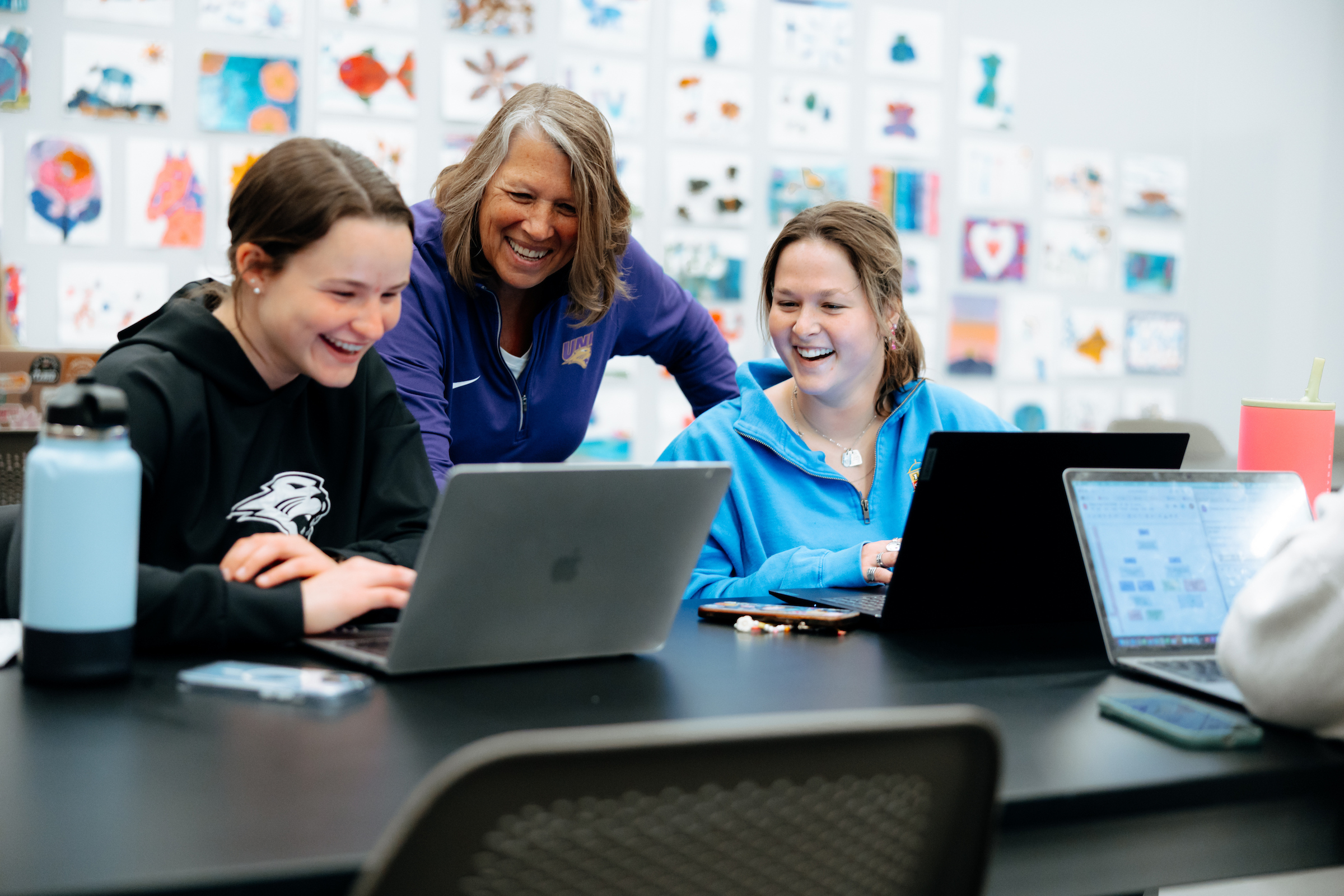 Several people gather around a large table in a classroom or collaborative learning space, working on laptop computers. One person wearing a purple UNI quarter-zip stands and leans over the table while interacting with two seated participants who are using laptops. Water bottles, smartphones, and other personal items are placed on the tabletop. In the foreground, additional laptops and a blurred chair are visible. The background wall is covered with numerous colorful drawings, paintings, and student-created