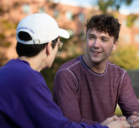 Two people sitting outdoors and talking. One wears a white cap and purple sweatshirt, and the other wears a maroon long-sleeve shirt, smiling as they converse.