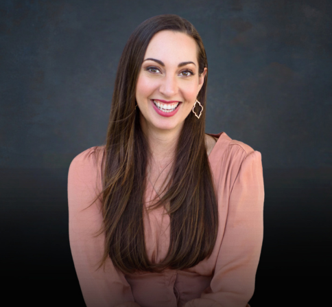 Studio headshot of a smiling woman (Vanessa Van Edwards) with long dark brown hair, wearing a soft pink blouse and geometric drop earrings. She is seated against a dark, textured backdrop and faces the camera with a friendly expression.
