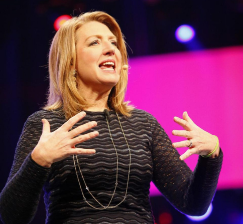 A woman with shoulder-length blonde hair is speaking on stage in front of bright, colorful lights and a large pink screen. She is wearing a dark, long-sleeved textured top and a long necklace. She is gesturing with both hands raised near her chest, appearing animated and engaged as she speaks.