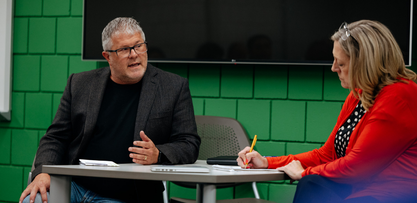 Two professionals are seated at a small meeting table in a conference room. A gray-haired man wearing glasses, a dark blazer, and a black shirt is speaking and gesturing with one hand while looking toward a woman seated beside him. The woman, wearing a red cardigan over a black-and-white polka dot blouse, is looking down and writing notes with a yellow pencil. Papers, a notebook, and a tablet are on the table. A large wall-mounted screen and a green brick wall are visible in the background.