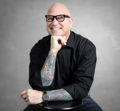 Portrait of a smiling bald man (Todd Dewett) wearing black-framed glasses and a black button-down shirt, seated on a stool against a light gray background. He rests his chin on his hand, showing colorful tattoo sleeves on both forearms and a wristwatch on his left wrist.