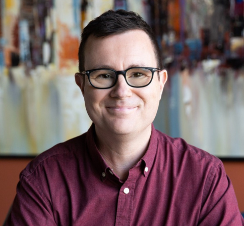 A person with short dark hair and black-framed glasses is smiling at the camera. They are wearing a maroon button-up shirt and sitting in front of a blurred, colorful abstract painting.
