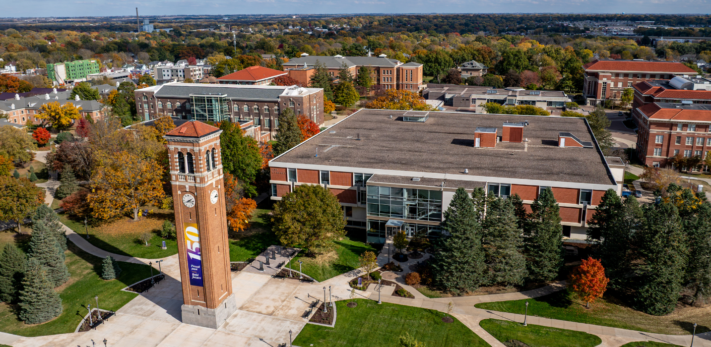Aerial view of a university campus during autumn, featuring a tall brick clock tower in a central plaza surrounded by sidewalks, lawns, and mature trees with colorful fall foliage. Several large red-brick academic buildings and parking areas extend across the campus, with a wooded landscape and city skyline visible in the distance under a clear blue sky. Visible text on a banner attached to the clock tower includes a large “150” and the words “Proud Past, Bold Future.”