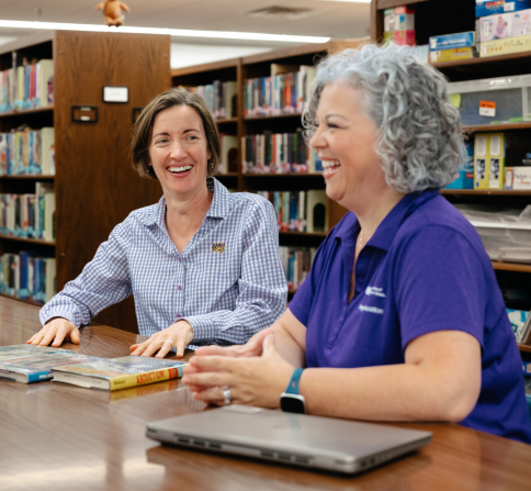 Two people sit at a wooden table in a library surrounded by bookshelves filled with books and educational materials. Several books are spread across the table, and a closed laptop sits in the foreground. One person wears a checkered button-down shirt and the other wears a purple polo shirt and a smartwatch. The library setting includes shelving, storage bins, and learning resources visible in the background.