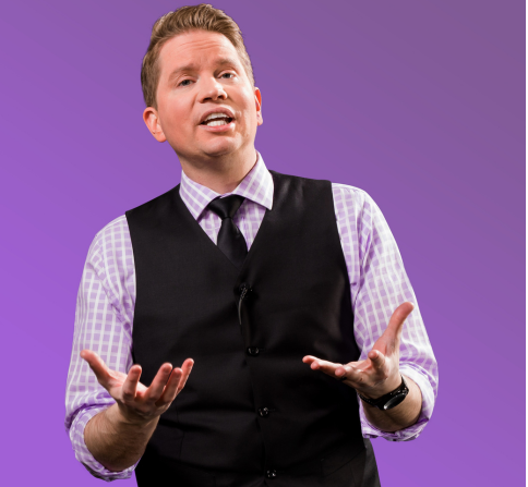 A person stands against a solid purple background, speaking with both hands raised in a gesturing position. The person is wearing a black vest over a light purple checkered dress shirt with a dark tie, along with a wristwatch. The image is cropped from about the waist up and appears to be a professional promotional or presentation photo. The clean background and studio lighting keep the focus on the speaker.