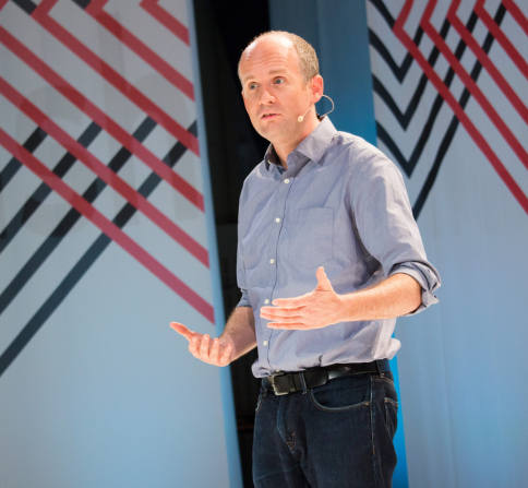 A person stands on a stage giving a presentation while wearing a headset microphone, a long-sleeved button-down shirt, jeans, and a belt. The person is positioned in front of large geometric backdrop panels featuring diagonal red, black, and white lines. The speaker is gesturing with both hands while addressing an audience. Bright stage lighting illuminates the scene, and the background includes modern event or conference-style decor.