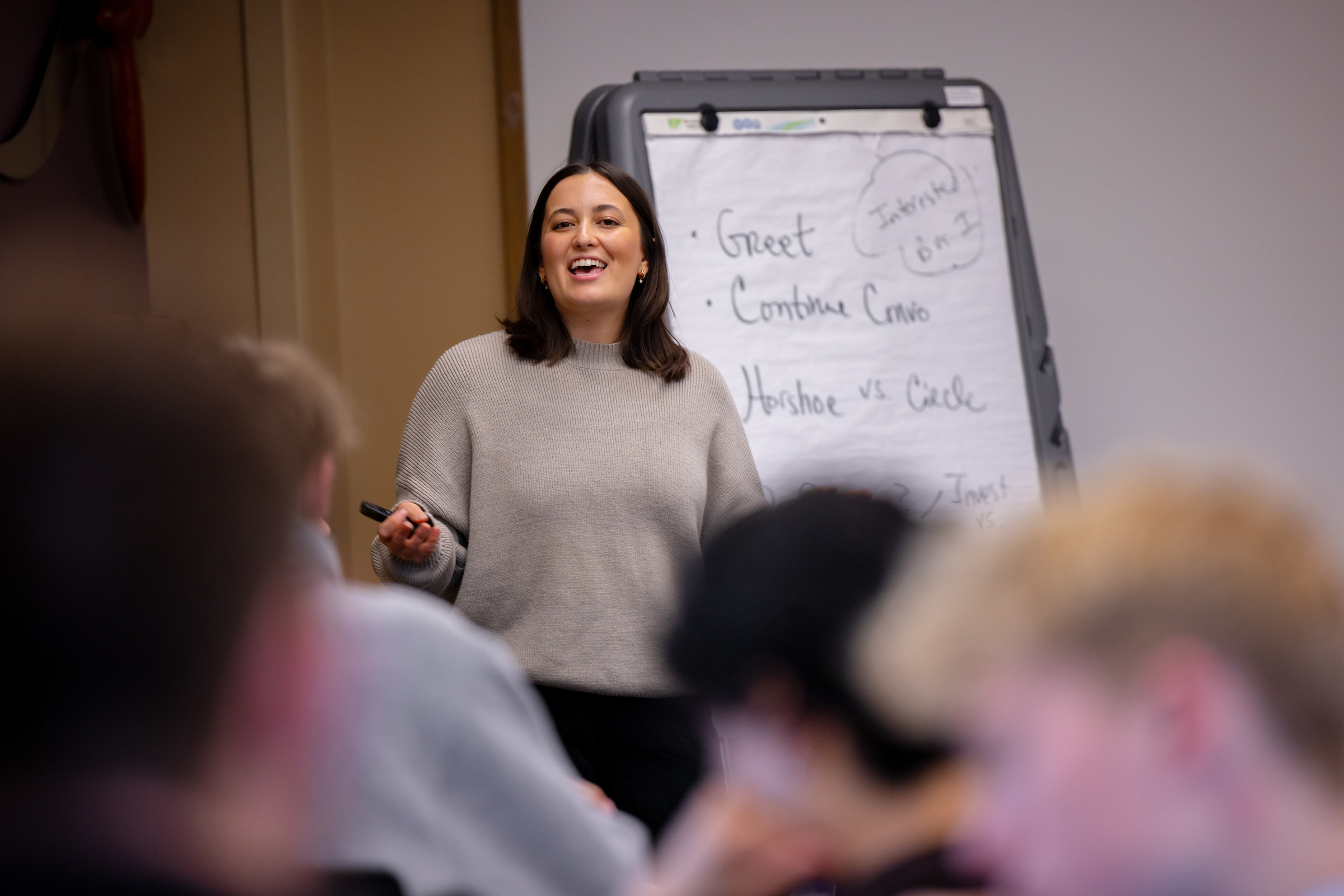 A person stands at the front of a meeting or training room delivering a presentation to a seated audience. The presenter wears a light-colored sweater and holds a presentation remote while speaking beside a flip chart easel. Handwritten notes on the flip chart include topics such as “Greet,” “Continue Conv.,” and “Horseshoe vs Circle.” Several audience members are visible in the foreground, out of focus, facing the presenter. The indoor setting appears to be a workshop, training session, or professional dev